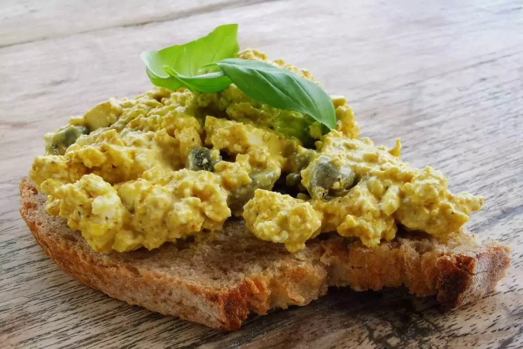 Gele tofu spread met curry op een een volkoren boterham, afgewerkt met basilcumblaadjes.