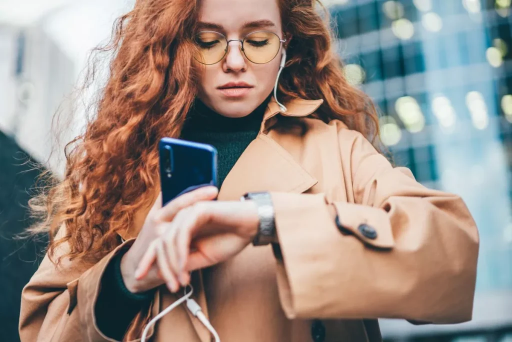 Mooi vrouw met krullen kijkt op haar klok terwijl ze naar muziek luistert en haar gsm vasthoudt.